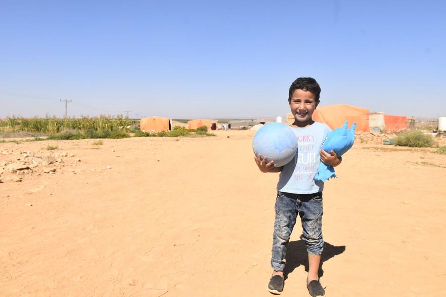A smiling boy holds a blue soccer ball and a stuffed toy